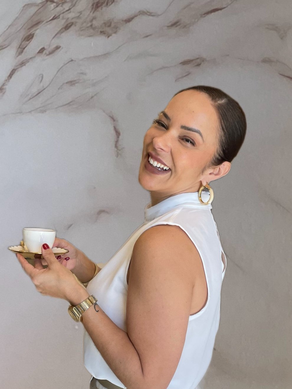 Dra. Ana Paula Neves sorrindo, de blusa branca, segurando uma xícara de café em frente a uma parede de mármore